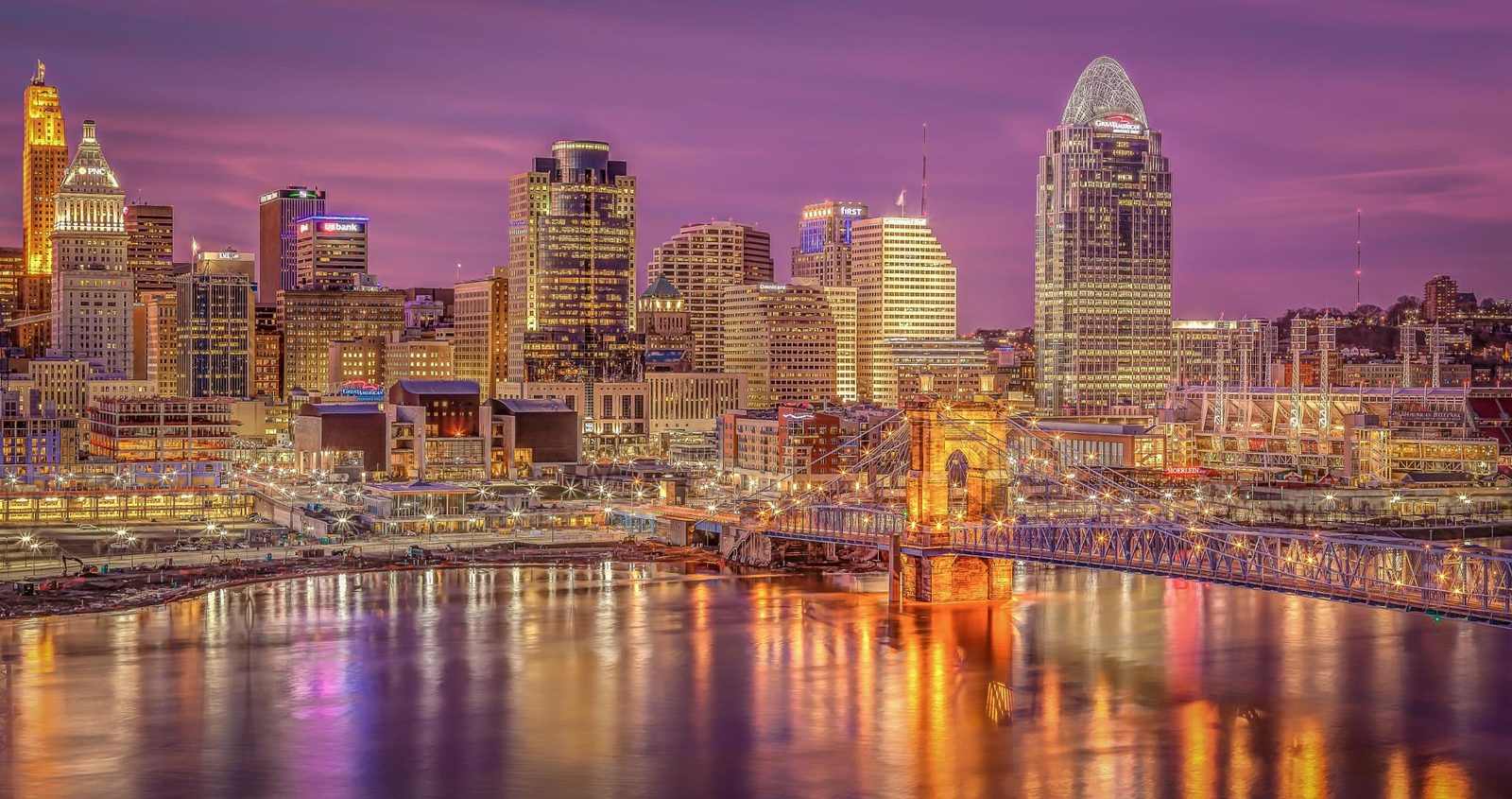 Cincinnati, Ohio downtown skyline at dusk reflecting in the Ohio River, with the John A. Roebling Suspension Bridge illuminated in the foreground